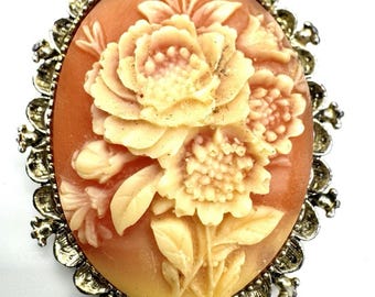 May include: An oval brooch featuring a carved cream-colored floral relief set against a warm peach-toned background. The central floral design is surrounded by an ornate, scalloped metallic frame with a textured, antiqued silver-tone finish.