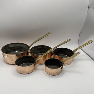 May include: A set of five graduated copper saucepans with polished brass handles. Each pan features a round, metallic copper body with a dark, weathered interior lining and a long, slender brass handle attached to the side with rivets.