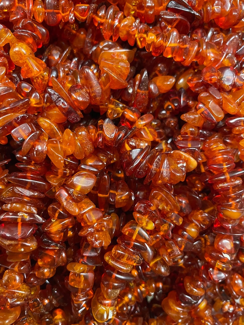 May include: Close-up of a collection of amber gemstone beads. The beads are a warm, translucent orange-brown color, with irregular shapes and sizes. The beads are strung together, creating a textured and organic appearance.