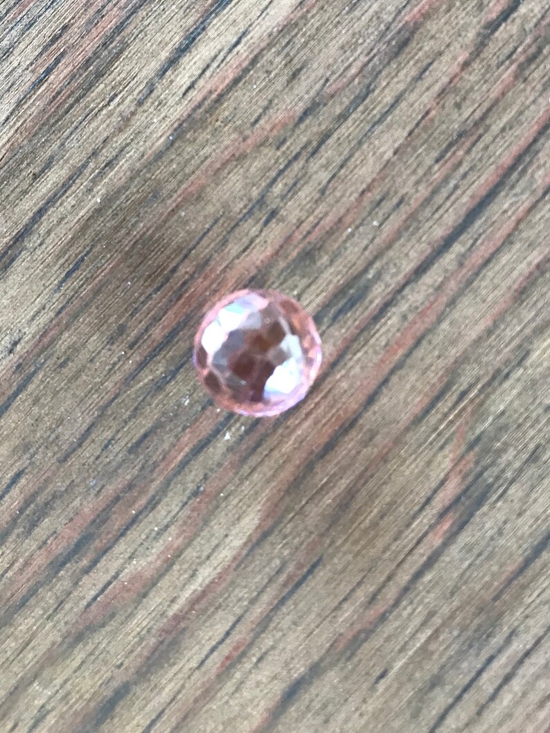 Puede incluir: Una sola piedra preciosa rosa facetada sobre una superficie de madera.