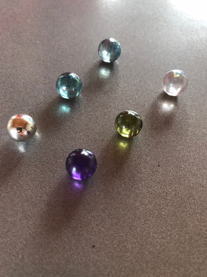 Puede incluir: Seis canicas de vidrio de varios colores, incluyendo azul, verde, morado y transparente, sobre una superficie gris.