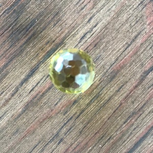 Puede incluir: Una sola piedra preciosa redonda, facetada y amarilla sobre una superficie de madera.
