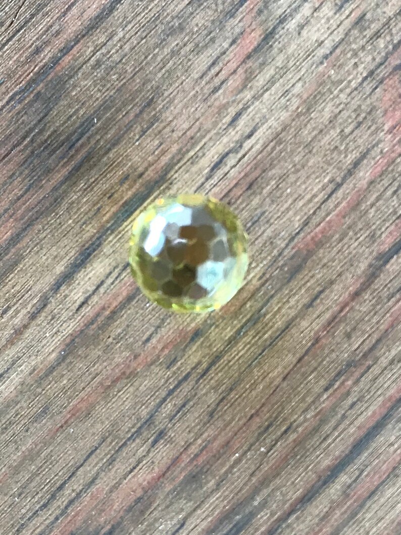 Puede incluir: Una sola piedra preciosa redonda, facetada y amarilla sobre una superficie de madera.