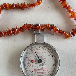 May include: A strand of polished amber beads, in shades of orange and brown, is laid across a silver ring gauge. The gauge, a "MM Gauge and Weight Estimator" by A. Leveridge, displays measurements and is marked "Renewed to Year 1994."