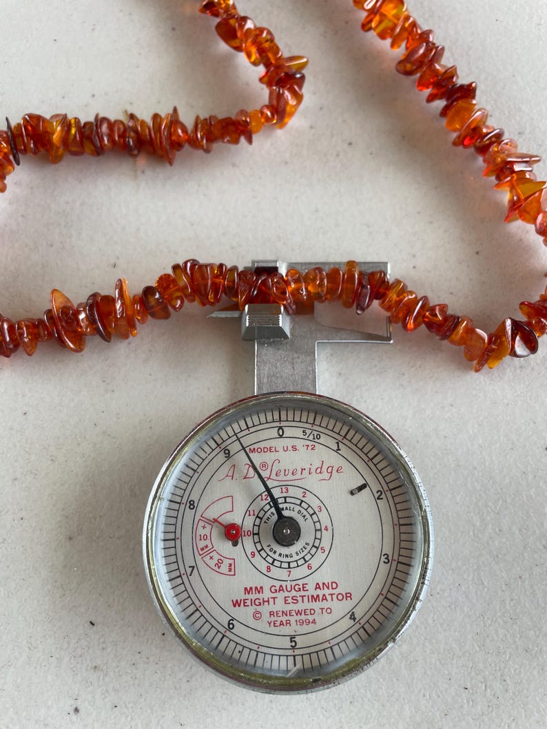 May include: A strand of polished amber beads, in shades of orange and brown, is laid across a silver ring gauge. The gauge, a "MM Gauge and Weight Estimator" by A. Leveridge, displays measurements and is marked "Renewed to Year 1994."