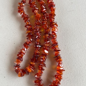 May include: A necklace made of polished amber stones. The amber stones are a warm, translucent orange-brown color and are irregularly shaped. The necklace is strung together, creating a flowing, organic design. The necklace is displayed against a neutral background.
