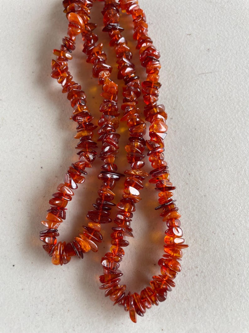 May include: A necklace made of polished amber stones. The amber stones are a warm, translucent orange-brown color and are irregularly shaped. The necklace is strung together, creating a flowing, organic design. The necklace is displayed against a neutral background.