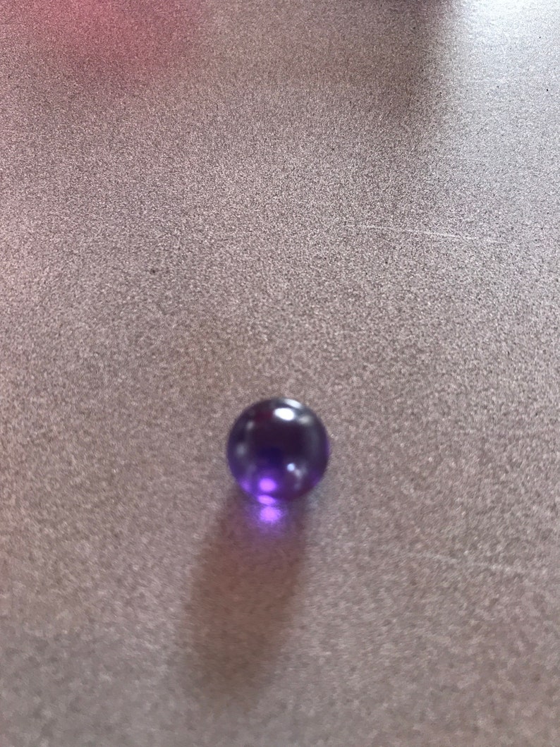Puede incluir: Un solo m&aacute;rmol de vidrio morado sobre una superficie gris texturizada.