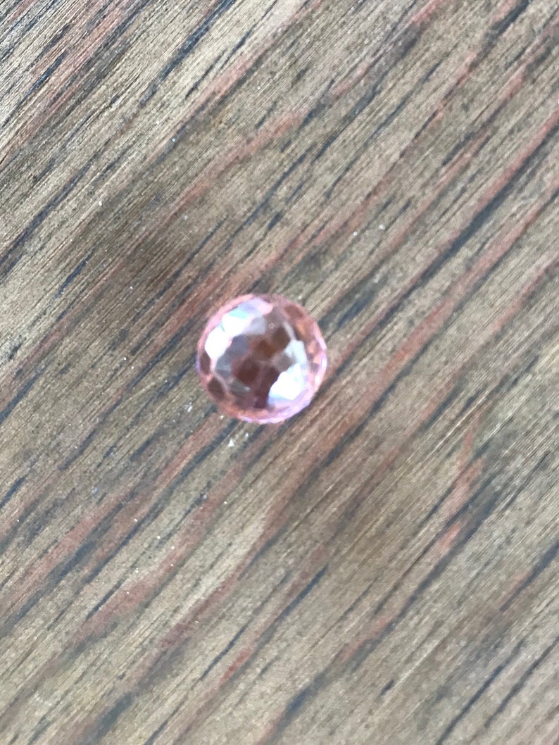 Puede incluir: Una sola piedra preciosa rosa facetada sobre una superficie de madera.