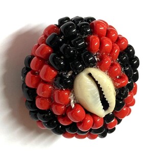 May include: A red and black beaded sphere with a white cowrie shell in the center. The beads are arranged in a spiral pattern.