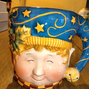 May include: A ceramic mug shaped like an elf's head with a blue and yellow hat. The mug has a whimsical design with stars and swirls. The elf has a friendly face with closed eyes and a smile.
