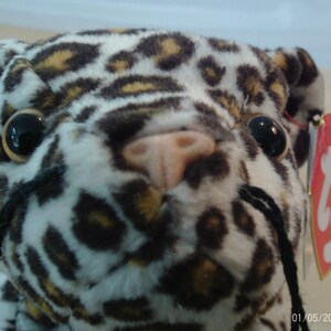 May include: A close-up of a leopard plush toy with a white, brown, and tan spotted pattern. The toy has large black eyes, a pink nose, and black whiskers. A red tag with gold text is attached.