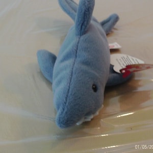 Puede incluir: Un juguete de peluche de tiburón azul claro con vientre blanco, ojos negros y pequeños dientes blancos. El tiburón tiene aletas y una etiqueta adjunta. El juguete está hecho de material suave.