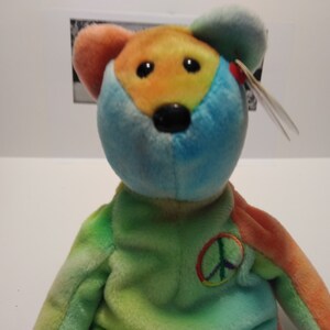 May include: A tie-dye plush toy bear with a peace sign on its chest. The bear is blue, green, orange, and yellow.