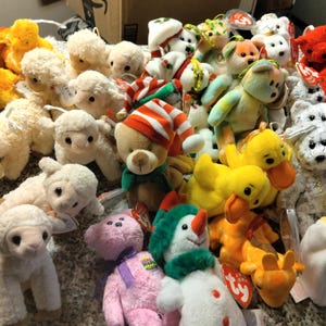 Puede incluir: Una colección de varios peluches, incluyendo corderos, patos, osos y muñecos de nieve. Los juguetes son de varios colores, incluyendo blanco, amarillo, marrón, rojo y rosa. Algunos juguetes tienen etiquetas.