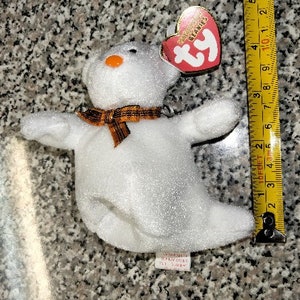 May include: White plush ghost beanie baby with an orange and brown ribbon around its neck. The beanie baby has a red Ty tag attached to its head that says "Ty Beanie Babies".