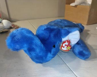 May include: A bright blue plush elephant toy with white inner ears and a small black eye. A heart-shaped red and yellow Ty tag is attached to the ear with a clear plastic fastener. The stuffed animal is lying down with its trunk curved upward.