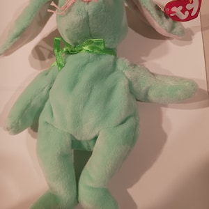 May include: A green plush bunny with long ears and a green bow. The bunny has a pink nose and black eyes. The bunny is wearing a Ty tag that says "Beanie Babies".