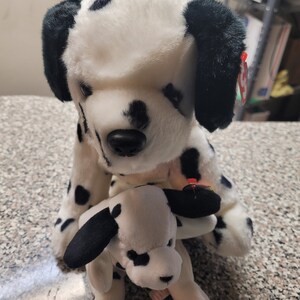 May include: A plush dalmatian dog toy with black spots on white fur, featuring black ears and a black nose. A smaller plush dalmatian puppy is positioned in front of the larger dog. Both toys have a red Ty tag attached to their ears.
