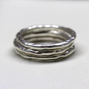May include: A set of four silver rings with a textured, organic design. The rings have a wavy, uneven surface, creating a unique and handcrafted look. The rings are displayed on a white background.