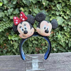 Puede incluir: Una diadema negra con dos caras de personajes con orejas y caras con cuentas negras. Un personaje tiene un lazo rojo con lunares blancos. La diadema tiene un exterior negro y un interior azul.