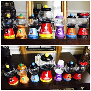 May include: Eight decorated glass candy jars with Mickey Mouse, Pluto, Pete, Goofy, Minnie Mouse, and Lucas Clubhouse themes. The jars are on a wooden table and have a red, yellow, blue, purple, and green base.