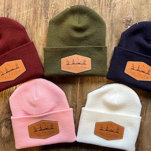 May include: Five beanies in various colors: burgundy, olive green, navy blue, pink, and white. Each beanie has a brown leather patch with the text "Fay's Pet Hay" and a graphic of a rabbit and reeds. The beanies are displayed on a wooden surface.