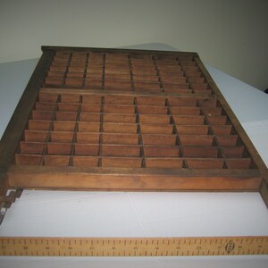 May include: A vintage wooden printing type case with a grid of compartments. The case is made of dark brown wood and has a worn, rustic appearance.