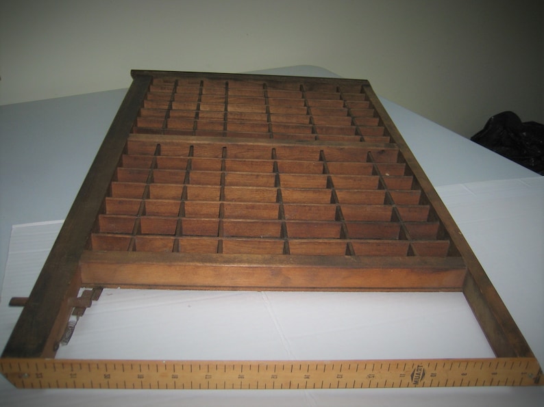 May include: A vintage wooden printing type case with a grid of compartments. The case is made of dark brown wood and has a worn, rustic appearance.