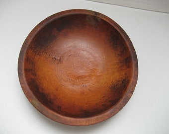 May include: A round wooden bowl with a deep basin and a thick, rounded rim. The surface of the bowl displays a rich, warm brown color with visible wood grain patterns and darker, weathered patches throughout the interior and exterior walls.