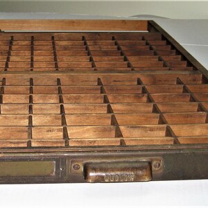 May include: A wooden printing type case with a brass drawer and a brass handle labeled "Ludlow". The case has a grid of compartments for storing individual pieces of type.