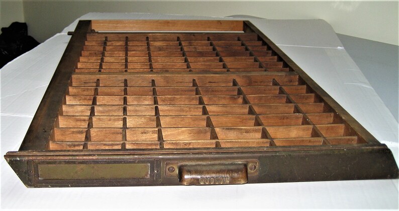 May include: A wooden printing type case with a brass drawer and a brass handle labeled "Ludlow". The case has a grid of compartments for storing individual pieces of type.