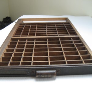 May include: A wooden printing tray with a metal drawer and a handle labeled "Ludlow". The tray has a grid of compartments for storing printing blocks.