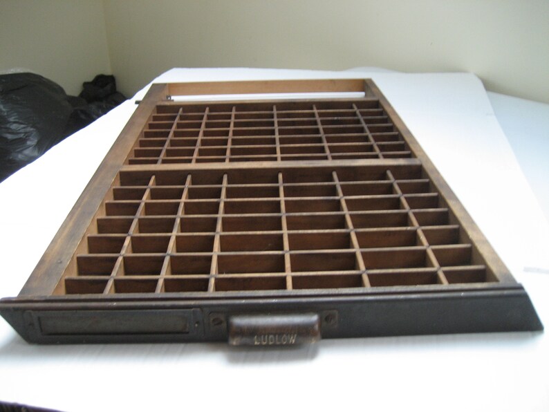 May include: A wooden printing tray with a metal drawer and a handle labeled "Ludlow". The tray has a grid of compartments for storing printing blocks.