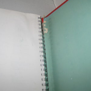 May include: A spiral-bound notebook with a red spine and a teal-colored cover. The notebook is open, revealing a white page on the left and a teal page on the right. The binding is made of silver metal.