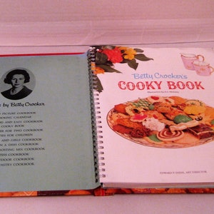 May include: An open vintage cookbook titled "Betty Crocker's Cooky Book" with a spiral binding, featuring a colorful illustration of cookies and a cup of tea. The left page lists other cookbooks by Betty Crocker.