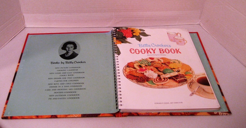 May include: An open vintage cookbook titled "Betty Crocker's Cooky Book" with a spiral binding, featuring a colorful illustration of cookies and a cup of tea. The left page lists other cookbooks by Betty Crocker.