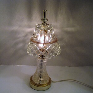 May include: A clear glass table lamp with a gold base and a diamond-patterned shade. The lamp is lit and casting a warm glow.