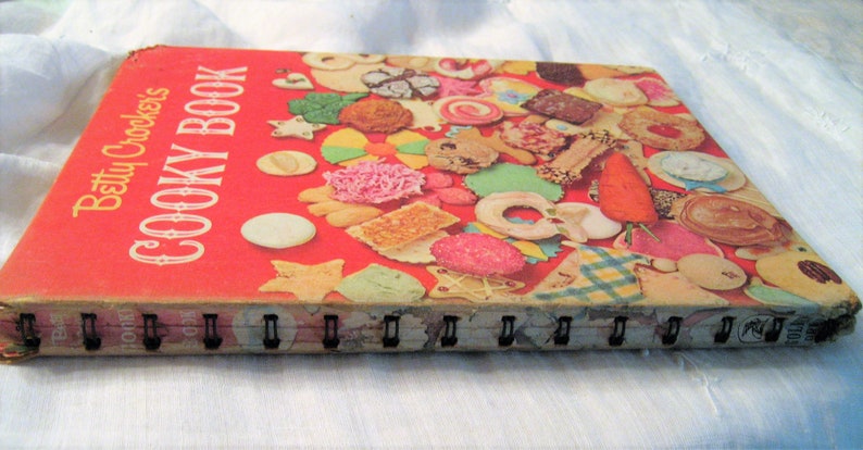 May include: Vintage "Betty Crocker's Cooky Book" with a red cover and spiral binding. The cover features a collage of colorful cookies and the title in white. The book's edges show wear, indicating its age.