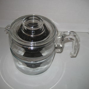 May include: A clear glass coffee percolator with a black metal filter and a glass lid. The percolator has a handle and a measurement line that says "6".