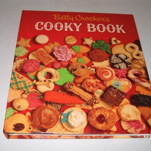 May include: A vintage cookbook titled "Betty Crocker's Cooky Book" with a red cover. The cover is adorned with a variety of colorful cookies, including stars, hearts, and gingerbread men. The book is open, showcasing the assortment of baked goods.