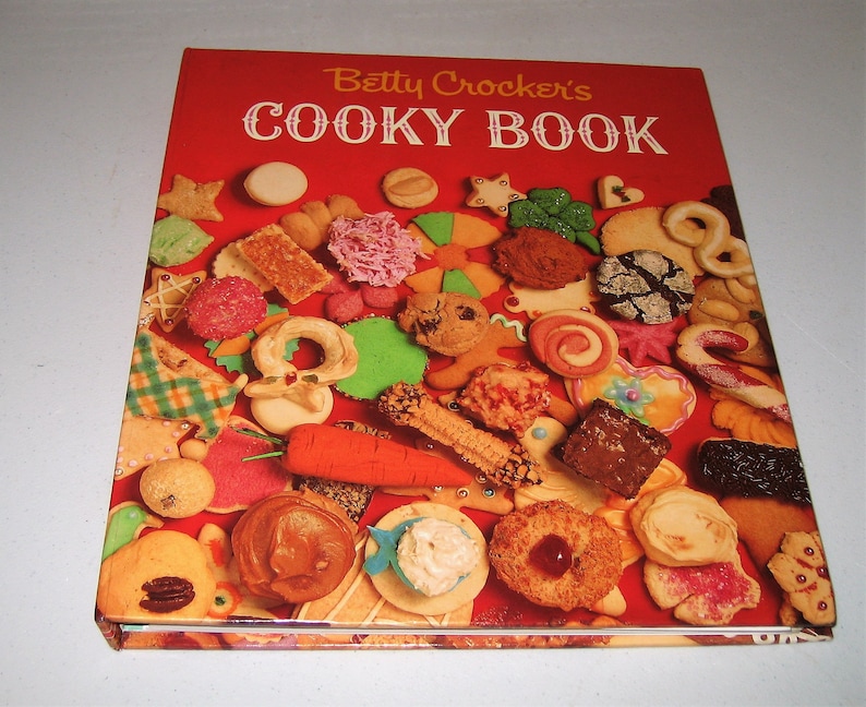 May include: A vintage cookbook titled "Betty Crocker's Cooky Book" with a red cover. The cover is adorned with a variety of colorful cookies, including stars, hearts, and gingerbread men. The book is open, showcasing the assortment of baked goods.