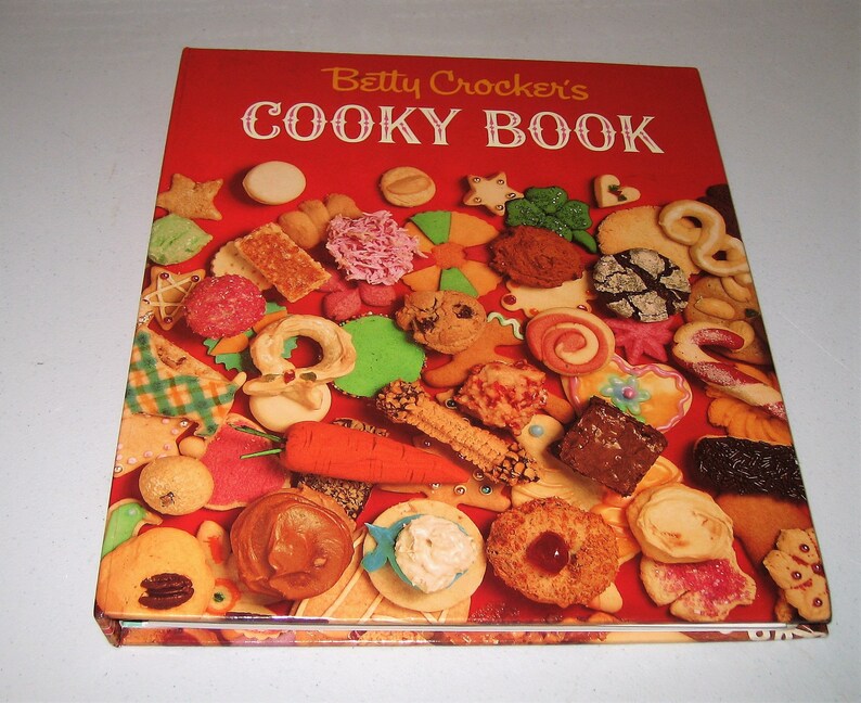 May include: A vintage cookbook titled "Betty Crocker's Cooky Book" with a red cover. The cover is adorned with a variety of colorful cookies, including stars, hearts, and gingerbread men. The cookies are arranged on a red background.