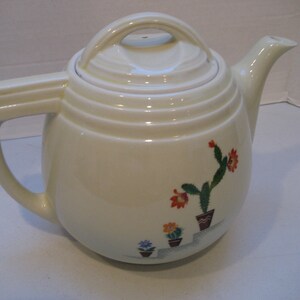 May include: White ceramic teapot with a lid and a handle. The teapot has a floral design with green cactus plants and orange, red, and blue flowers.