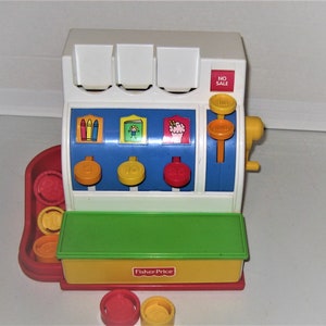 May include: A white, blue, green, and yellow plastic toy cash register by Fisher-Price. It features three colorful buttons labeled 5, 10, and 25, a red No Sale button, a side crank, and a coin tray containing several plastic coins in red and yellow.