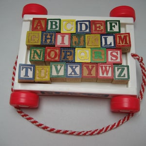 May include: A white wooden toy wagon with red wheels and a rope handle. The wagon is filled with colorful wooden blocks that spell out the alphabet.