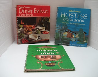 May include: Three vintage cookbooks with spiral binding. Titles include "Dinner for Two," "Hostess Cookbook," and "Dinner in a Dish." The covers feature illustrations of food and entertaining scenes. The books are in good condition, showing signs of age.