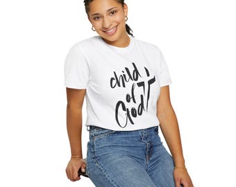 May include: A white t-shirt with the text "Child of God" in black script. The shirt is short-sleeved and worn by a person in blue jeans. The person is smiling and seated on a black stool.