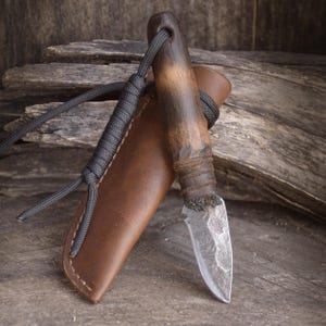 May include: A small fixed-blade knife with a textured, dark metal blade and a smooth, two-toned brown wooden handle. The knife is accompanied by a brown leather sheath featuring a dark gray paracord lanyard wrapped around the handle and sheath.