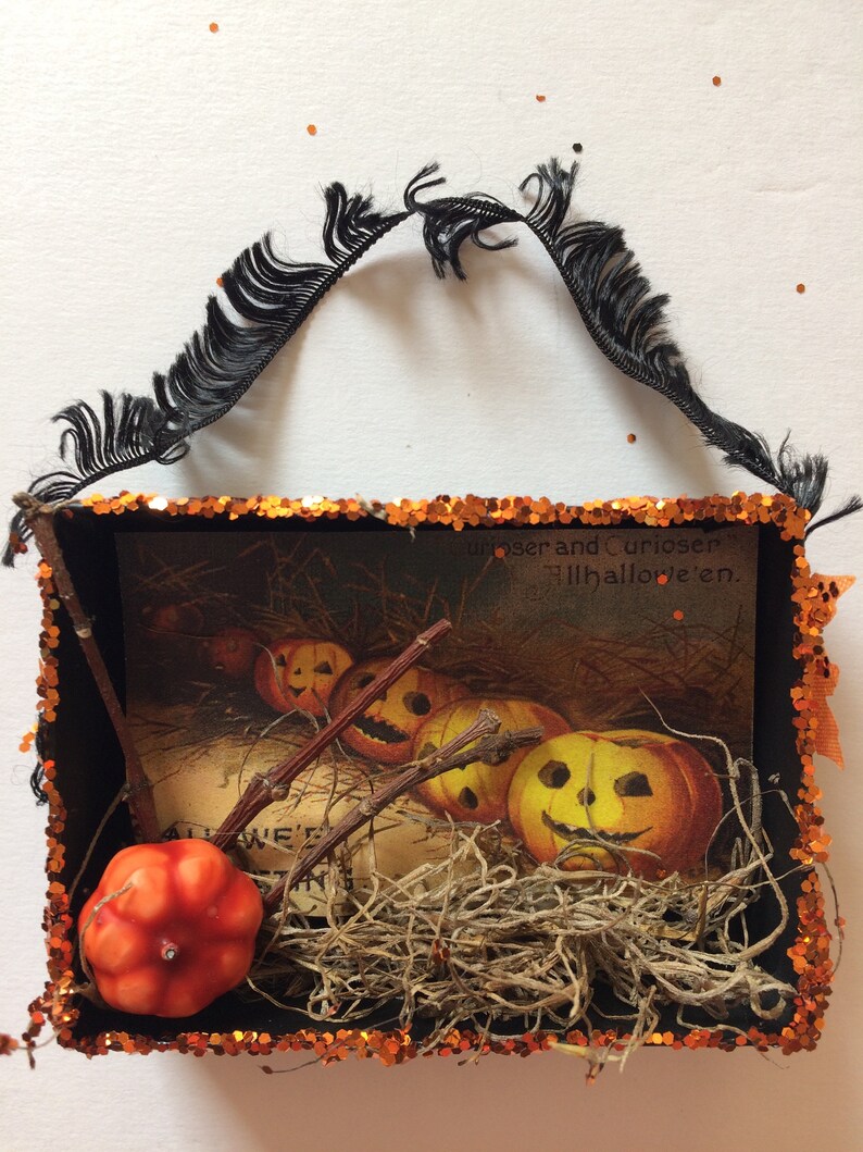 Pu&ograve; includere: Una scatola ombra a tema Halloween con dettagli arancioni e neri. Contiene un'illustrazione vintage di zucche intagliate, ramoscelli e muschio secco. La parte superiore ha una frangia nera e i bordi sono rifiniti con paillettes arancioni.